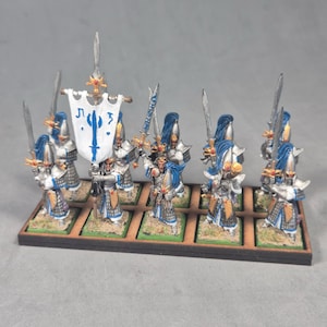 May include: Miniature figurines of warriors in silver armor with blue accents and long blue hair. Each figure holds a sword and stands on a small, green-covered base. One figure carries a banner with a blue design. The figurines are arranged on a wooden base.