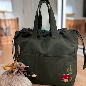 Könnte beinhalten: Dunkelgrüne Tragetasche mit zwei Vordertaschen und Kordelzugverschluss. Die Tasche hat ein gesticktes rotes Pilz- und grünes Blattdesign. Ein kleiner Blumenstrauß und ein Wollknäuel liegen neben der Tasche.