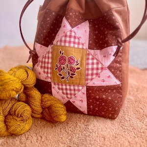 Peut inclure: Un sac marron à cordon de serrage avec un motif d'étoile en patchwork. L'étoile est faite de tissu à carreaux roses et floral, avec un carré central représentant une illustration florale. Une pelote de fil jaune est posée à côté du sac.