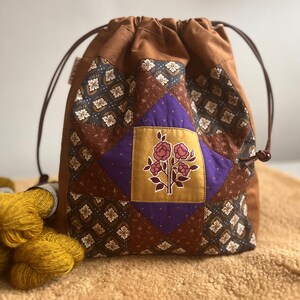May include: A brown drawstring bag with a patchwork design. The patchwork features floral and geometric patterns in shades of brown, purple, and gold. A floral motif is centered on the front. The bag is filled with yellow yarn.