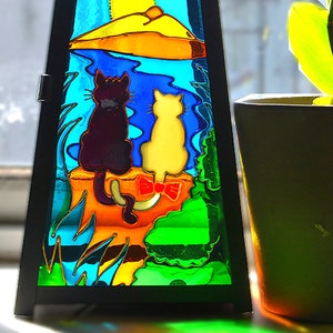 May include: A black metal lantern with a stained glass design featuring two cats and a sun. The lantern is sitting on a windowsill next to a potted pink orchid.
