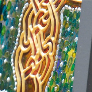 Op de afbeelding: Close-up van een mozaïek van gekleurd glas met een geel, oranje en rood ontwerp. Het ontwerp is omgeven door een rand van kleine, groene, blauwe en witte glasstukjes.