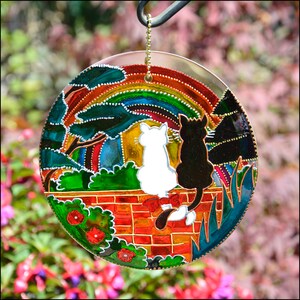 May include: A round, colorful stained glass suncatcher with a rainbow, two cats, and a brick wall. The cats are black and white and are sitting on the wall. The suncatcher is hanging from a chain.