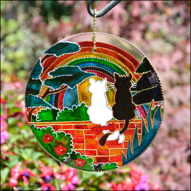 May include: A round, colorful stained glass suncatcher with a rainbow, two cats, and a brick wall. The cats are black and white and are sitting on the wall. The suncatcher is hanging from a chain.