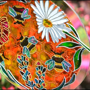 May include: A round, clear glass plate with a painted design of a honeycomb, bees, flowers, and leaves. The honeycomb is orange and yellow, the bees are black and yellow, the flowers are white and yellow, and the leaves are green.