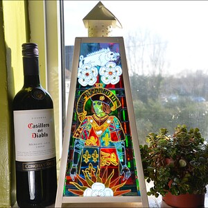 May include: A stained glass lamp with a triangular base and a colorful design featuring a knight in armor, a white boar, and flowers. The lamp is sitting on a windowsill next to a bottle of Casillero del Diablo Merlot wine.