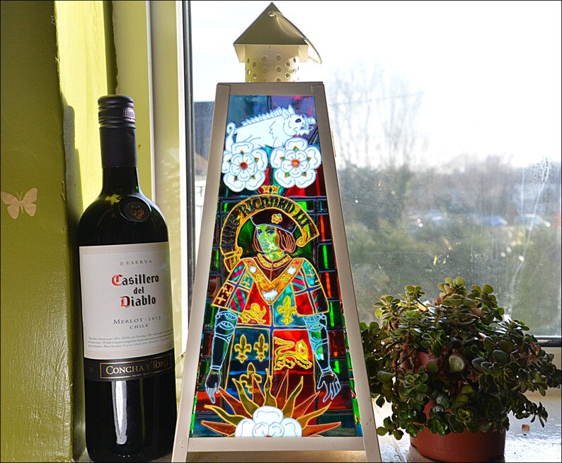 May include: A stained glass lamp with a triangular base and a colorful design featuring a knight in armor, a white boar, and flowers. The lamp is sitting on a windowsill next to a bottle of Casillero del Diablo Merlot wine.