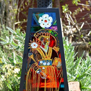 May include: A black metal lantern with a stained glass panel depicting a crowned figure in a red and gold robe. The figure is holding a staff and a book. The stained glass panel also features a white flower and a star.