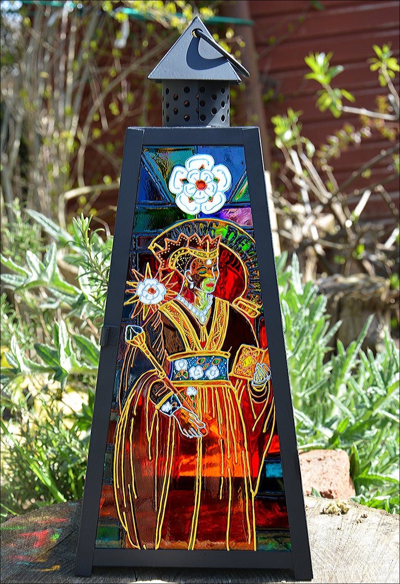 May include: A black metal lantern with a stained glass panel depicting a crowned figure in a red and gold robe. The figure is holding a staff and a book. The stained glass panel also features a white flower and a star.