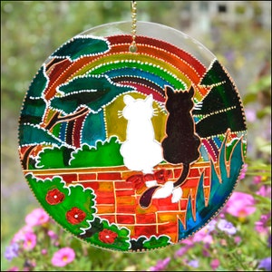 May include: A stained glass suncatcher with a rainbow, two cats, and a brick wall. The cats are black and white and are sitting on the wall. The suncatcher is round and has a gold chain for hanging.