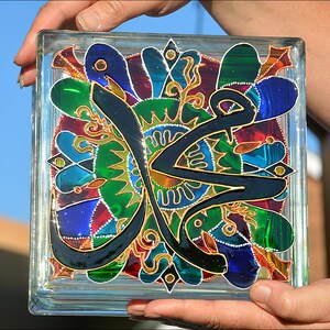 May include: A glass block with a colorful stained glass design featuring a black Arabic calligraphy inscription. The design includes green, blue, red, and gold accents.