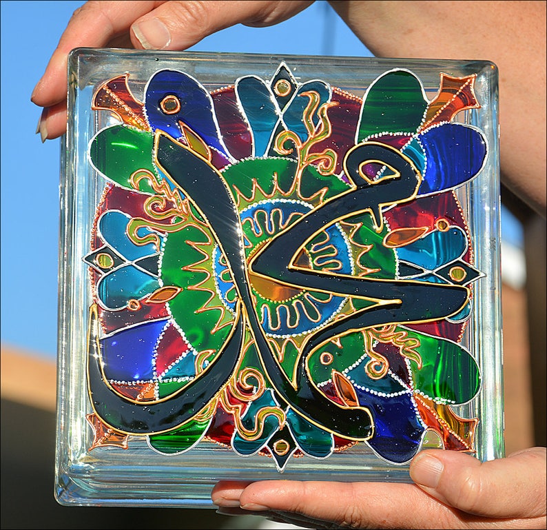 May include: A glass block with a colorful stained glass design featuring a black Arabic calligraphy inscription. The design includes green, blue, red, and gold accents.