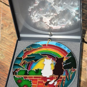 May include: A stained glass suncatcher featuring two cats sitting on a brick wall under a rainbow. The cats are facing forward and looking up at the rainbow. The suncatcher is in a black box with a gray foam lining.