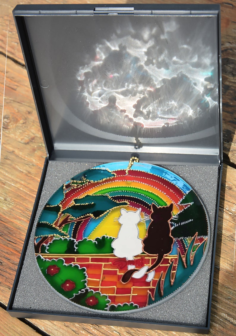 May include: A stained glass suncatcher featuring two cats sitting on a brick wall under a rainbow. The cats are facing forward and looking up at the rainbow. The suncatcher is in a black box with a gray foam lining.