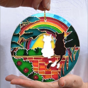 May include: A round stained glass suncatcher with a rainbow, two cats, and a brick wall. The cats are white and black and are sitting on the wall with their backs to each other. The suncatcher is surrounded by green foliage.