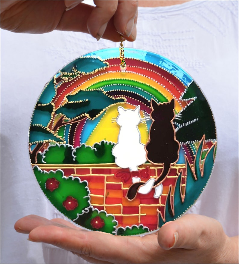 May include: A round stained glass suncatcher with a rainbow, two cats, and a brick wall. The cats are white and black and are sitting on the wall with their backs to each other. The suncatcher is surrounded by green foliage.