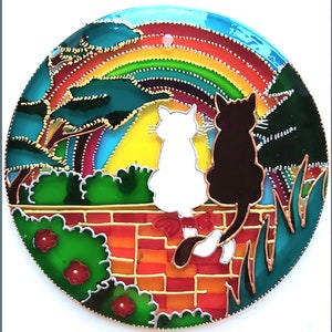 May include: A stained glass artwork depicting two cats sitting on a brick wall, looking at a rainbow in the sky. The cats are white and black, and the background is a colorful landscape with trees and grass.