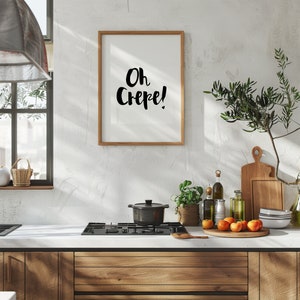 May include: A rectangular wall art print in a light wood frame featuring the black handwritten text Oh Crepe! on a white background. The print is displayed above a kitchen counter with a black stovetop, a pot, fresh fruit, and wooden boards.