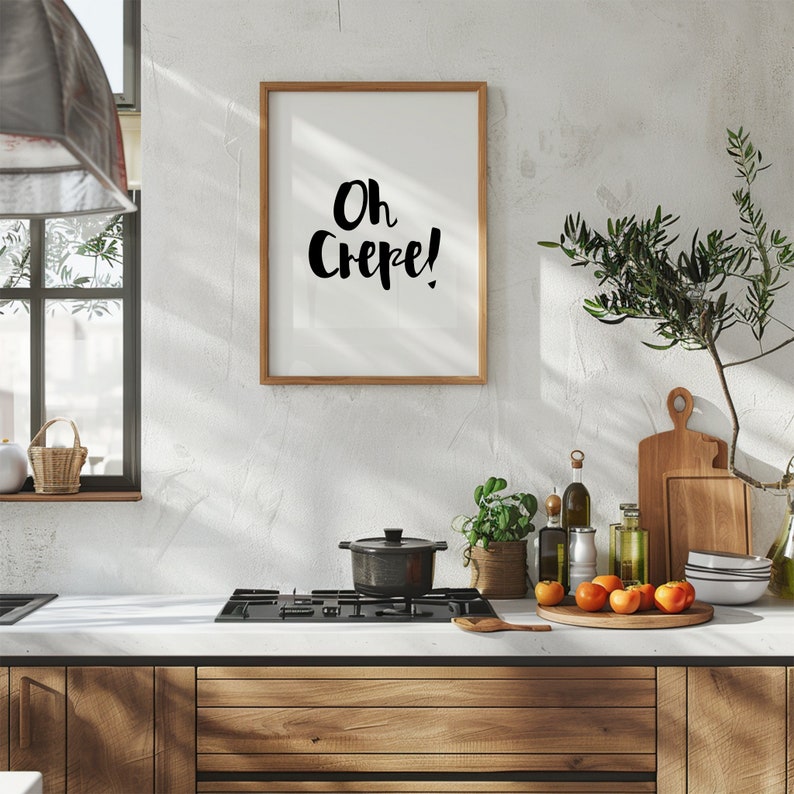 May include: A rectangular wall art print in a light wood frame featuring the black handwritten text Oh Crepe! on a white background. The print is displayed above a kitchen counter with a black stovetop, a pot, fresh fruit, and wooden boards.
