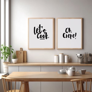 May include: Two framed wall art prints with light wood frames featuring black text on a white background. The left print displays the words "let's Cook" in a stylized script, while the right print displays the words "Oh Crepe!" in black script.