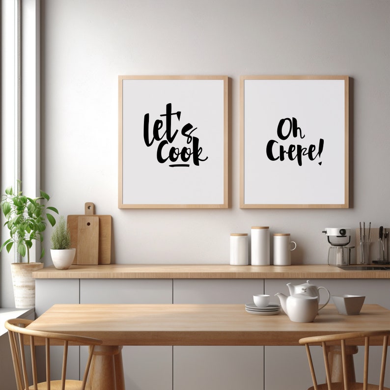 May include: Two framed wall art prints with light wood frames featuring black text on a white background. The left print displays the words "let's Cook" in a stylized script, while the right print displays the words "Oh Crepe!" in black script.