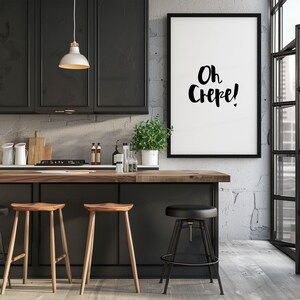 May include: A framed vertical wall art print featuring the black text Oh Crepe! in a stylized script font. The artwork is displayed in a kitchen setting with dark cabinetry, a wooden countertop, two light wood stools, and one black metal stool.