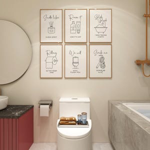 Six framed white prints with black bathroom-themed phrases and illustrations hang on a light-colored wall above a white toilet and a pink vanity.