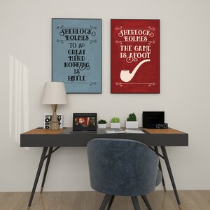 Puede incluir: Dos carteles enmarcados con citas de Sherlock Holmes cuelgan sobre un escritorio. Uno es azul claro con la cita "Para una gran mente nada es peque&ntilde;o". El otro es rojo con un gr&aacute;fico de pipa y la cita "El juego ha comenzado".