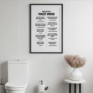 A framed white poster with black text listing toilet jokes hangs on a white ribbed wall above a white toilet and a vase with pink flowers.