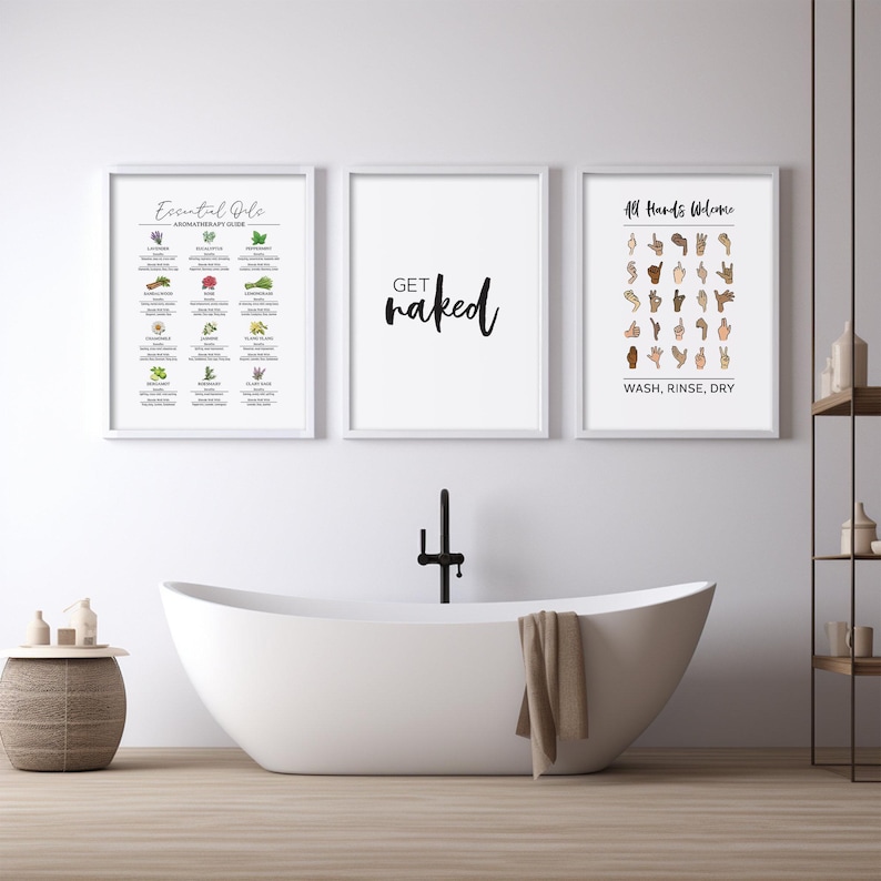 Three framed white prints hang above a white freestanding bathtub: one with essential oils, one saying "get naked," and one with ASL for "All Hands Welcome."