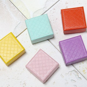 May include: Five small, square gift boxes in yellow, pale blue, red, purple, and pink are shown from above. Each box has a quilted pattern and a white dotted border. They are arranged on a light-coloured, textured surface.