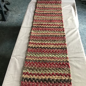 May include: A woven table runner with a multi-colored striped pattern in shades of red, brown, green, and beige. The runner is made of a thick, textured fabric and has a fringed edge.
