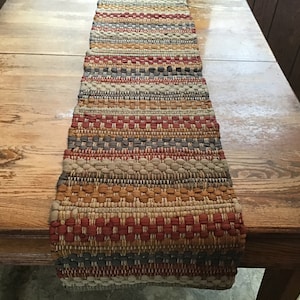 May include: A woven table runner with a multi-colored striped pattern in shades of brown, beige, gray, and red. The runner is made of a textured fabric and has a slightly frayed edge.