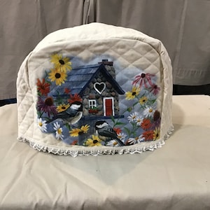 May include: A beige quilted toaster cover with a white lace trim. The cover features a painted design of a stone cottage with a red door and a heart-shaped window. Two birds are perched on branches with yellow, white, and pink flowers.