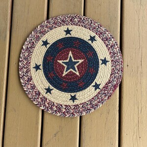May include: A round braided placemat featuring a concentric design with a central dark blue star on a maroon background, surrounded by a cream-colored ring with navy blue stars, and an outer border of interwoven red, white, and blue fibers.