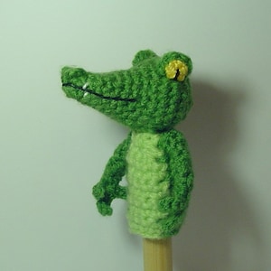 May include: Green crocheted alligator puppet with a yellow eye. The puppet is on a wooden stick.