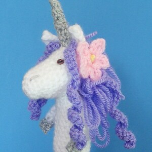 May include: White crocheted unicorn puppet with a purple mane and a pink flower on its head. The unicorn has a gray horn and is wearing a gray crocheted nose.