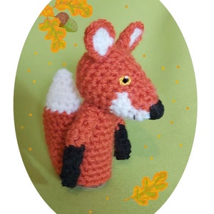 May include: A crocheted fox puppet with orange fur, white accents, and black feet. The puppet is posed with its tail curled up and its head tilted to the side. The background is a green oval with yellow dots and orange leaves.