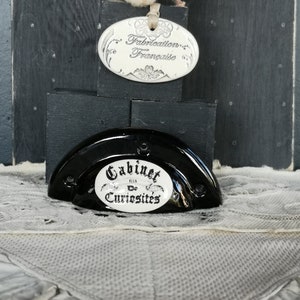 Peut inclure: Poignée de porte de cabinet en céramique noire avec une plaque ovale blanche sur laquelle est inscrit "Cabinet De Curiosites". Une plaque ovale blanche avec le texte "Fabrication Française" est également visible sur l'image.