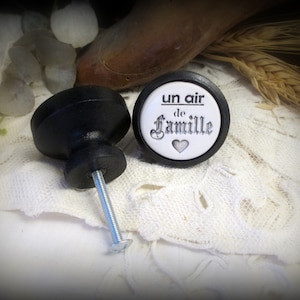 May include: Black and white ceramic knob with the text "un air de famille" and a small heart. The knob is attached to a black wooden base with a silver screw.