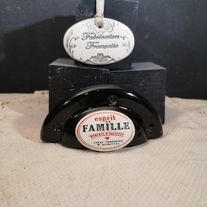 Może przedstawiać: Biały ceramiczny owalny ornament z czarnym tekstem "Fabrication Française" i białą wstążką. Czarny ceramiczny ornament z białym ceramicznym kółkiem, na którym widnieje napis "Esprit de Famille Moments à Partager Amour Tendresse & Affection" z czerwonym sercem.