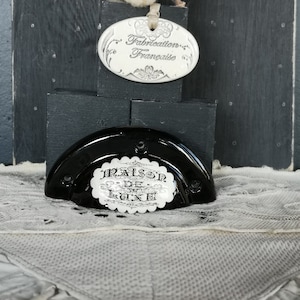 May include: A black and white ceramic door knob with the text "Maison de Luxe" in a circle. The knob is on a black wooden surface with a white cotton flower and a white oval tag with the text "Fabrication Française".