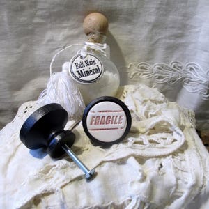 May include: Two white ceramic knobs with black bases. One knob has the word "FRAGILE" printed in red. The other knob has a white tag with black text that reads "Fait Main Mineral".
