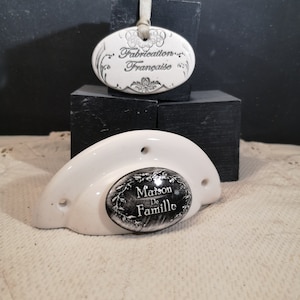 Peut inclure: Poignée de porte en céramique blanche avec un insert ovale noir qui indique "Maison de Famille". Une étiquette ovale blanche séparée indique "Fabrication Française".