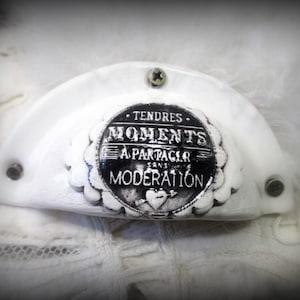 Puede incluir: Adorno de cerámica blanca con un diseño en blanco y negro. El adorno tiene el texto "Tendres Moments a Partager sans Moderation".