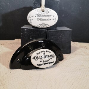May include: A black ceramic cabinet knob with a white oval ceramic label that reads "Prenez votre déjeuner LE CACAO PAYRAUD en feuilles PRODUIT SANS RIVAL +" and a smaller white oval ceramic label that reads "Fabrication Française".