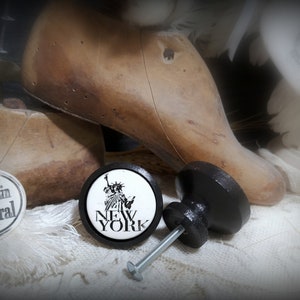 May include: Black and white ceramic knob with a picture of the Statue of Liberty and the words "New York" on it. The knob is attached to a black metal screw. The knob is sitting on a white fabric surface.