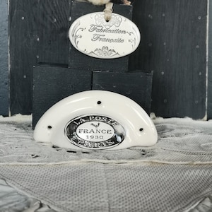 May include: A white ceramic half-moon shaped dish with a silver circular emblem that reads "La Poste France 1930 Paris" with a rooster graphic in the centre. The dish has three holes on the top and bottom. The dish is sitting on a white lace tablecloth.