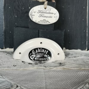 Peut inclure: Bouton de porte en céramique blanche avec une étiquette ovale noire et argentée qui indique "Cabinet de Curiosites 1713".