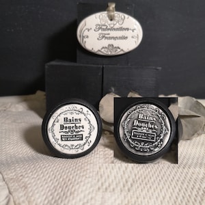 May include: Two white ceramic knobs with black trim and the text "Bains Douches" in a decorative font. The knobs are on a white textured surface. The text on the knobs reads "Bains Douches, Savon de Marseille et d'Olive"